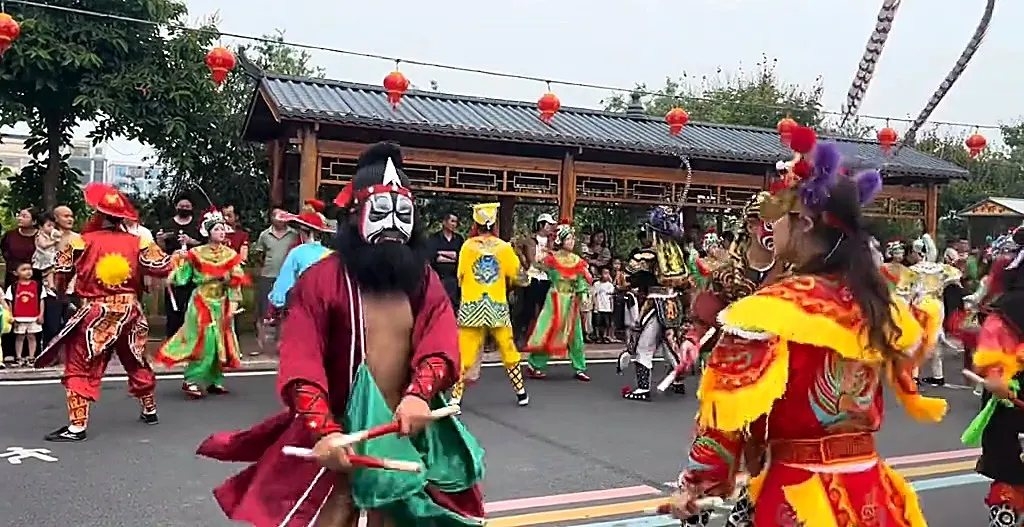 一名黑臉長鬚、紅袍的隊員雙手各執一根木槌站在街心,身後是彩繪長廊與紅燈籠