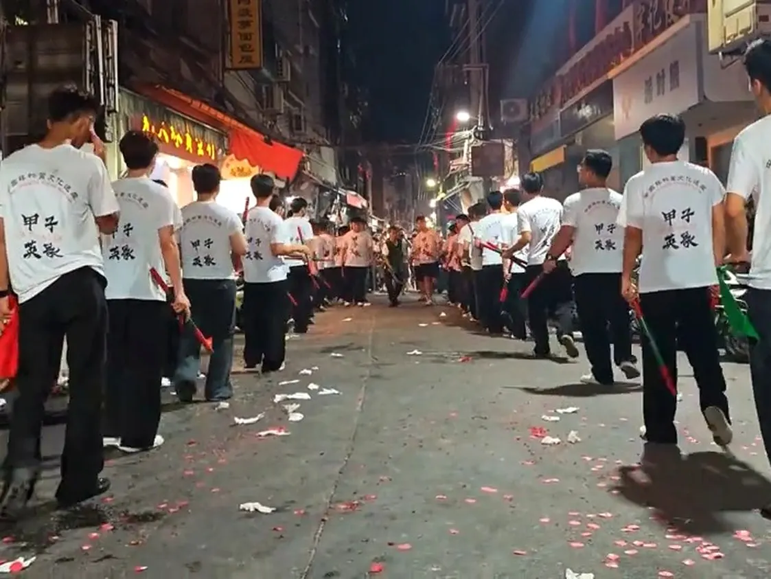 白 T 恤背後印著甲子英歌，隊伍踩著滿地紅紙往前走