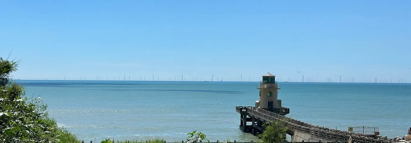 海邊小燈塔立在石砌防波堤盡頭,遠處海平線排著一列海上風機