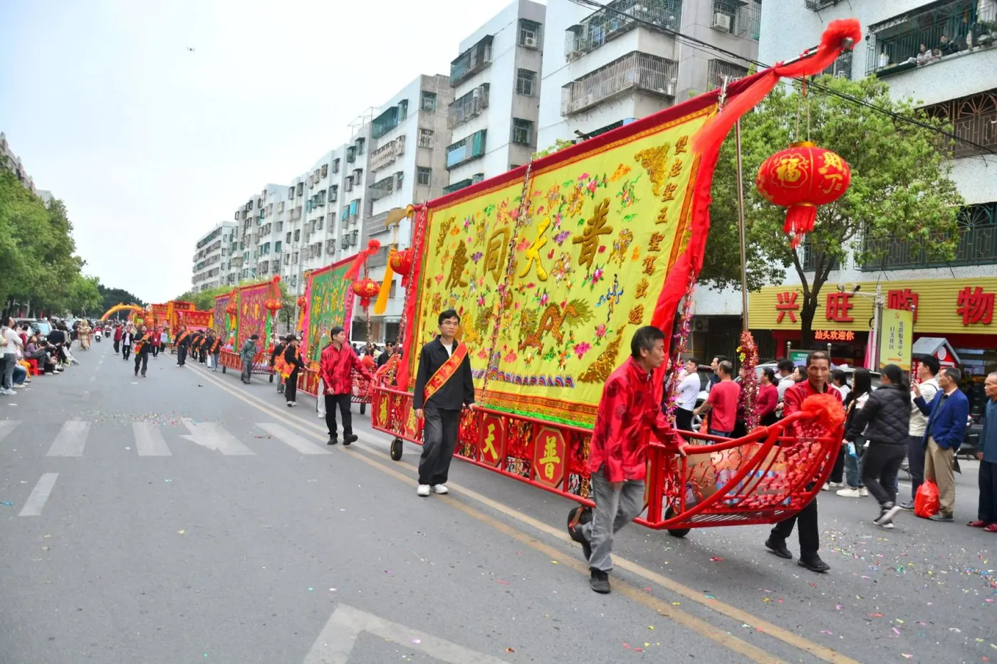 巡游队伍抬着绣有「普天同庆」的大幅标旗沿街行进,两旁是商铺与观众
