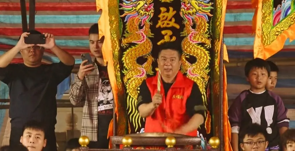 鼓師在繡雙龍與「鼓」字的黑幡前舉槌，鼓擱在四角帶金球的木架上