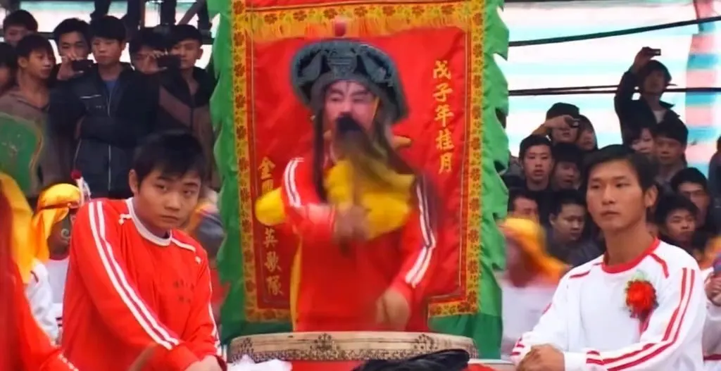 勾臉掛須的鼓手站在大鼓後，雙手舉槌，身後紅幡寫「戊子年桂月」