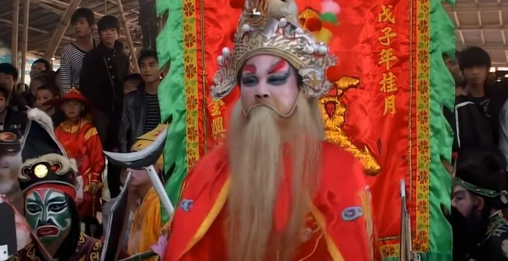 勾臉白底紅紋，掛白鬚、戴金冠，身後是繡金的紅幡