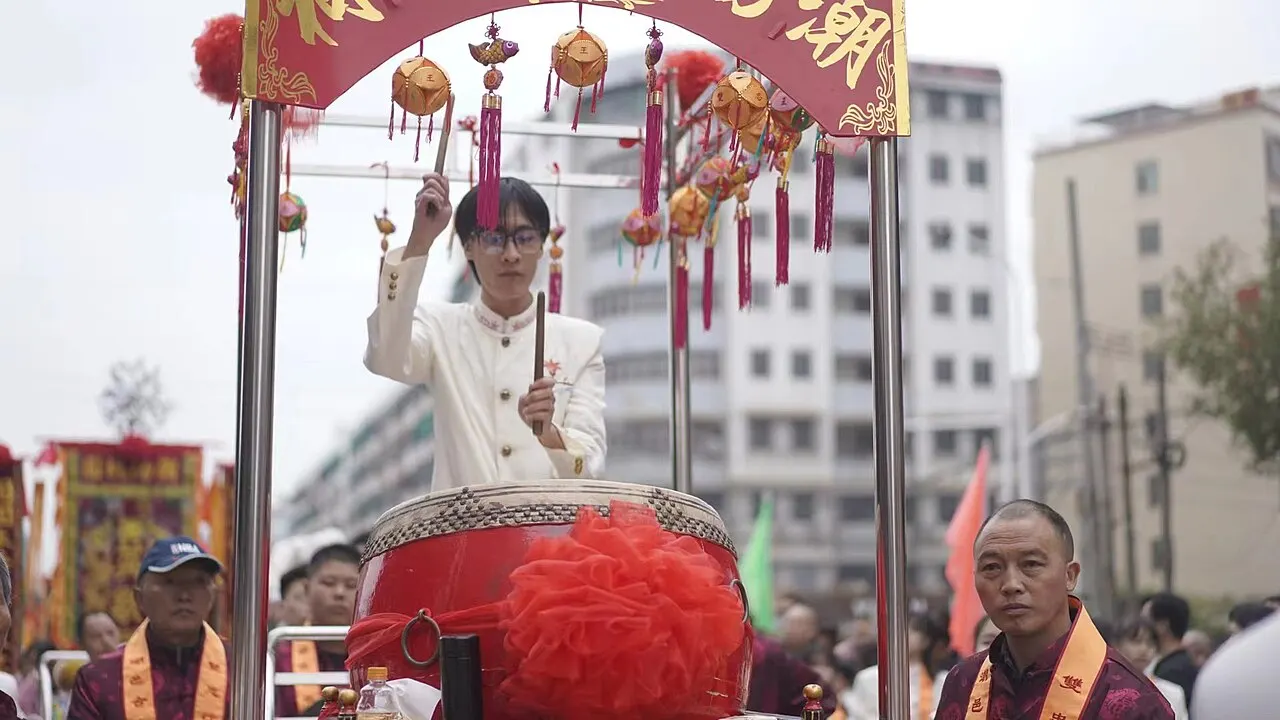 营老爷队伍中，一人立于鼓亭击鼓，两侧人员佩红绶带